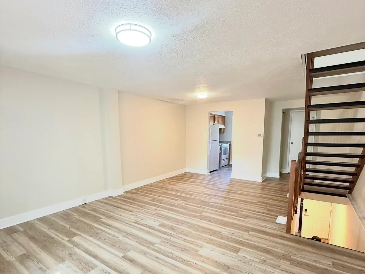 After: living room with a staircase and kitchen doorway, going from salmon walls and worn carpet to warm neutral paint and new plank flooring