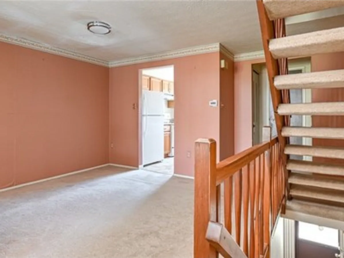 Before: living room with a staircase and kitchen doorway, going from salmon walls and worn carpet to warm neutral paint and new plank flooring